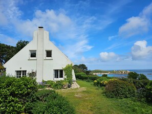 A white house with a steep roof, surrounded by lush greenery and a view of the sea. - Maison de Vacances à Louer sur L'ile Callot - Ker Douara (Carantec)