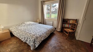 A bedroom with a bed, a nightstand, a chair, a bookshelf, and a window with curtains. - Maison de Vacances à Louer sur L'ile Callot - Ker Douara (Carantec)