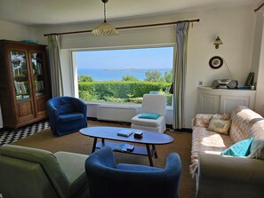 Ein Wohnzimmer mit Meerblick, ein blauer Sessel, ein weißer Sessel, ein Sofa, eine Holzkommode und ein Tisch mit Büchern.