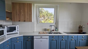 Una cocina con gabinetes azules, un lavavajillas blanco, un horno empotrado y un ventanal con vista a árboles.
