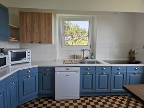 A kitchen with blue cabinets, a white dishwasher, a built-in oven, and a window with a view of trees. - Maison de Vacances à Louer sur L'ile Callot - Ker Douara (Carantec)