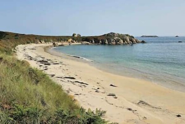 Uma praia com areal arenoso, águas cristalinas e afloramentos rochosos ao longe.