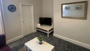 Un salon avec une table basse blanche, un téléviseur posé sur un meuble, une porte et un tableau encadré au mur.