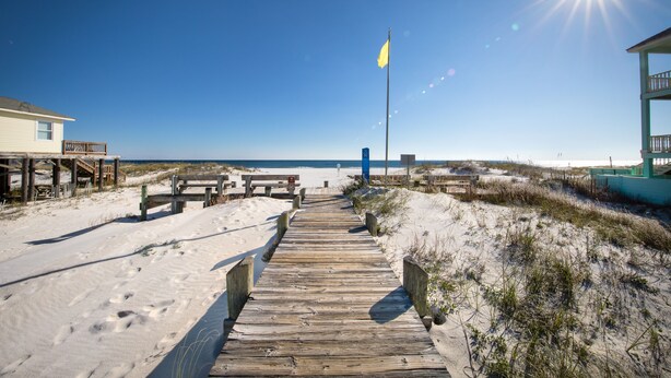 Une promenade en bois menant à une plage, avec un drapeau, des dunes de sable et des installations de plage.