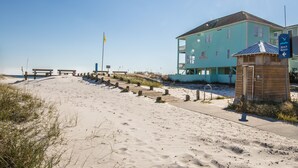 Un bâtiment situé au bord de la plage, avec un chemin dégagé menant à la plage, un poste de surveillance des sauveteurs et un panneau indiquant l’accès à la plage.