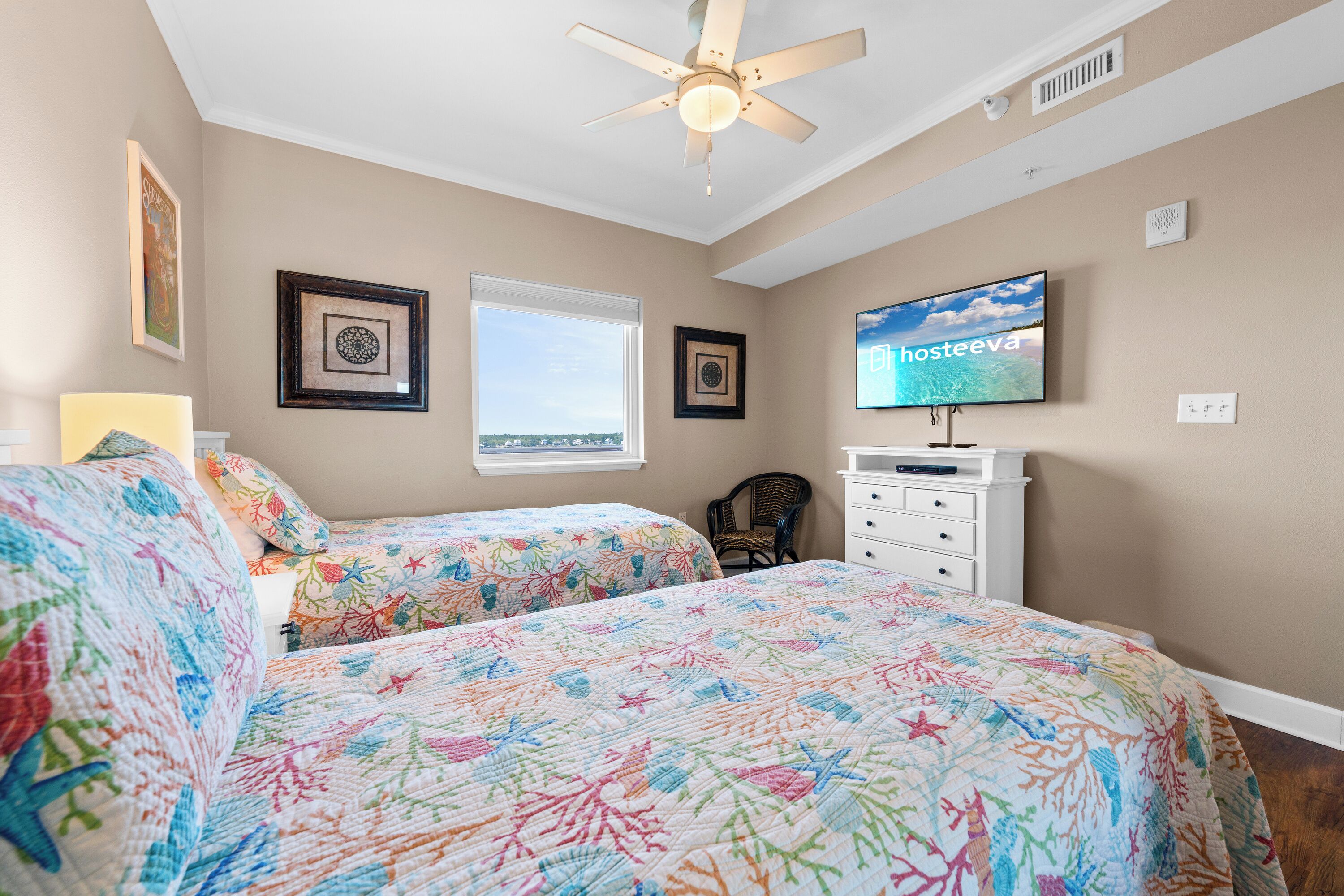 A bedroom with two beds, a ceiling fan, a television, and framed artwork on the walls.