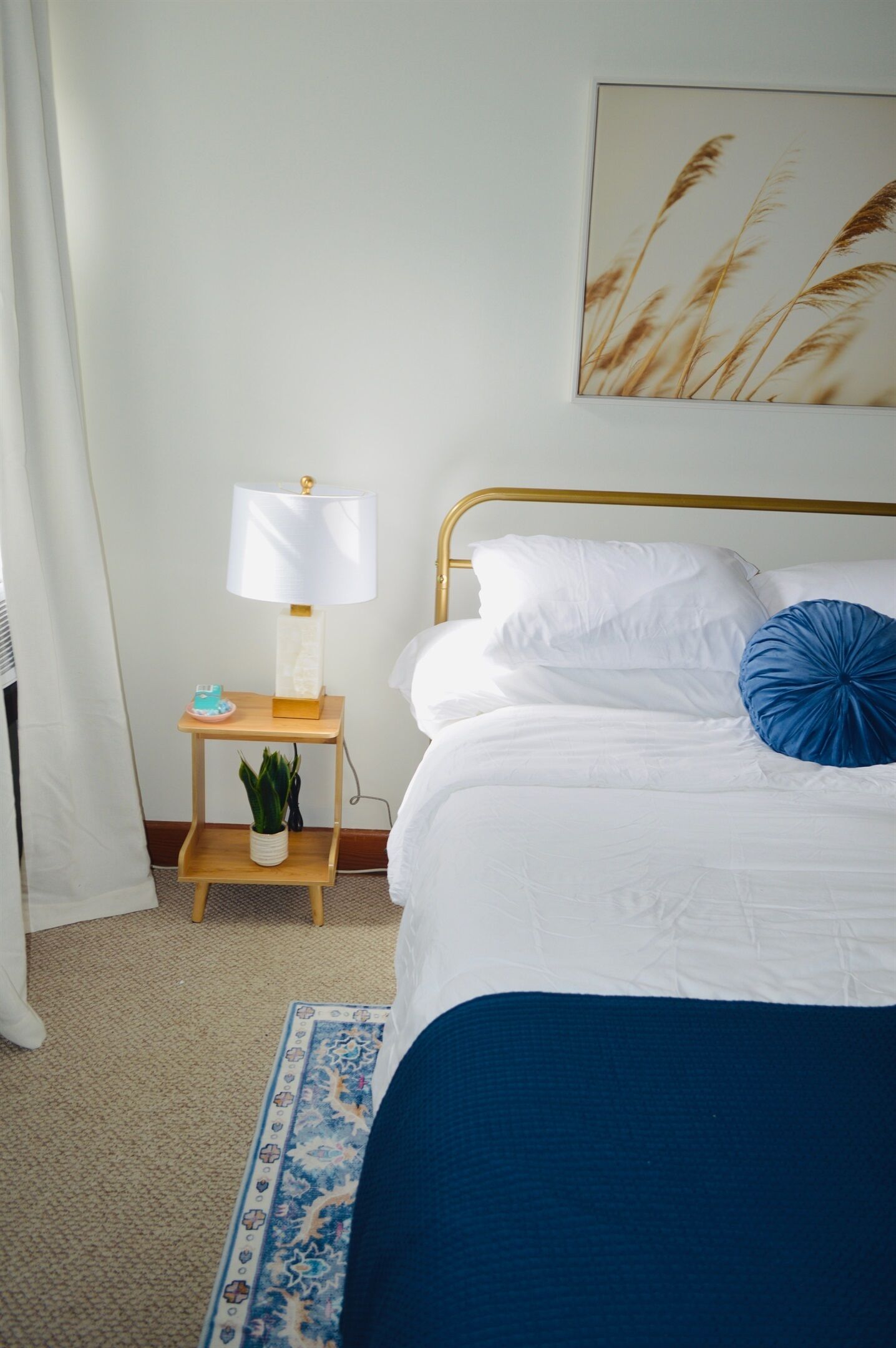 Uma cama bem arrumada com roupa de cama branca e azul, uma mesa de cabeceira com abajur, uma planta pequena e um quadro na parede.