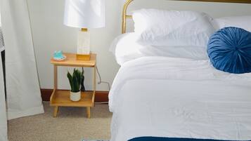 Uma cama bem arrumada com roupa de cama branca e azul, uma mesa de cabeceira com abajur, uma planta pequena e um quadro na parede.
