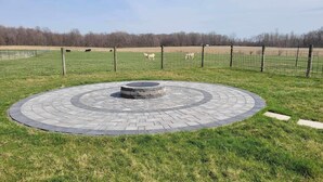 Ein kreisförmiger gepflasterter Bereich mit einer zentralen, brunnenartigen Struktur, umgeben von einer Wiese und einem Zaun.