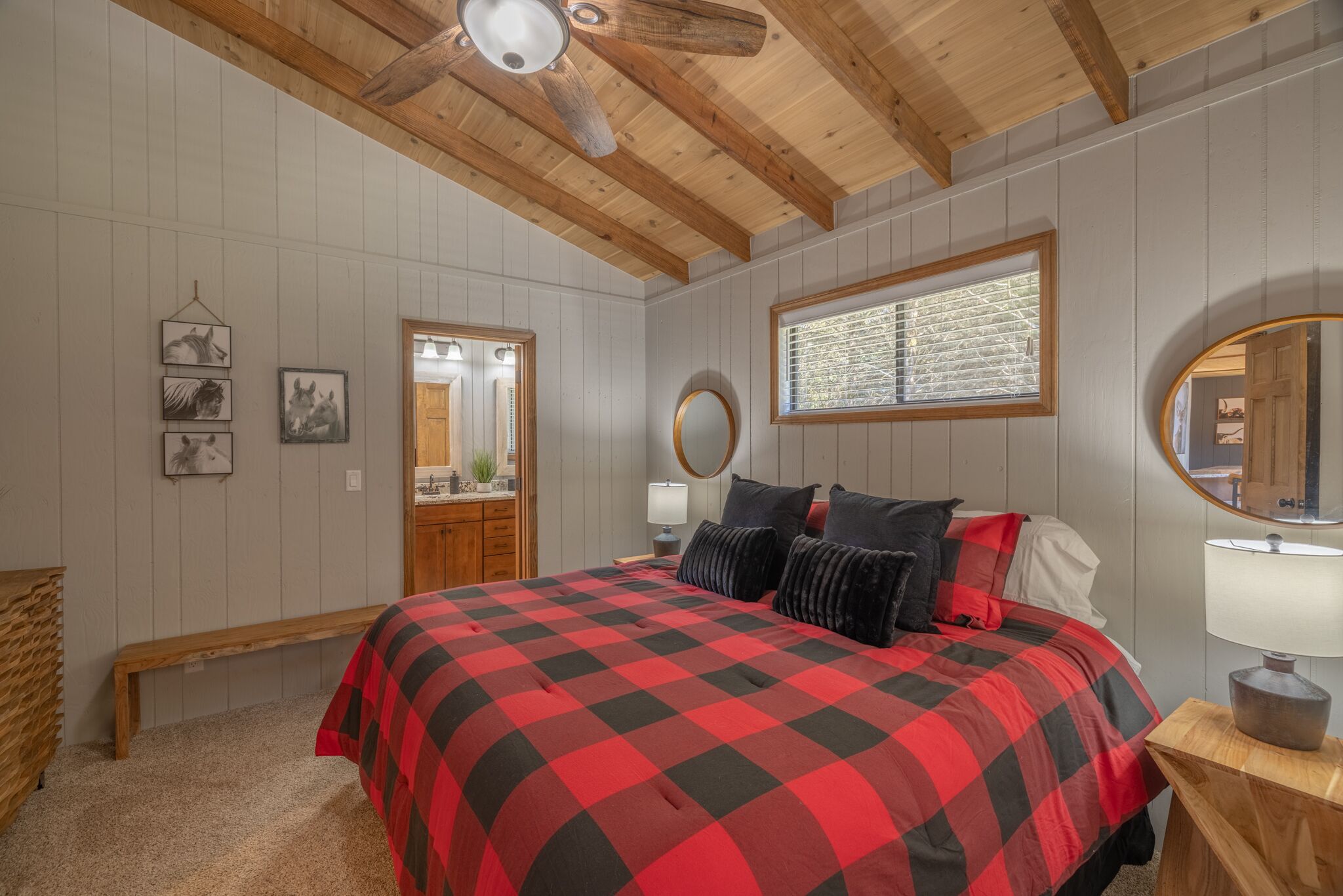 Un dormitorio con una cama grande, vigas de madera en el techo, una ventana con persianas, un espejo y una mesita de noche con una lámpara.