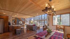 A spacious living area with a wooden ceiling, a kitchen with wooden cabinets, a dining table with checkered placemats, and large windows offering a view of the outdoors. - White Tail Lodge by Innsbrook Vacations! (Innsbrook)