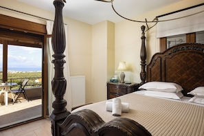 A bedroom with a four-poster bed, a view of the sea through a balcony, and a small table with chairs outside.
