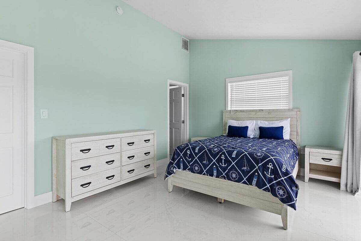 Une chambre à coucher comprenant un lit, une commode, une table de chevet et une fenêtre avec des stores.