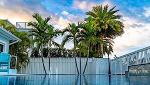 En swimmingpool med palmer, et hvidt hegn og et hus i baggrunden.