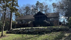 A two-story house with a stone retaining wall in front, surrounded by trees and a clear blue sky. - Cozy 3-bedroom lakefront cottage in beautiful Clemson, SC  (Clemson)