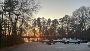 A parking lot with several vehicles, including a white SUV and a pickup truck, surrounded by trees and a sunset in the background. - Cozy 3-bedroom lakefront cottage in beautiful Clemson, SC  (Clemson)