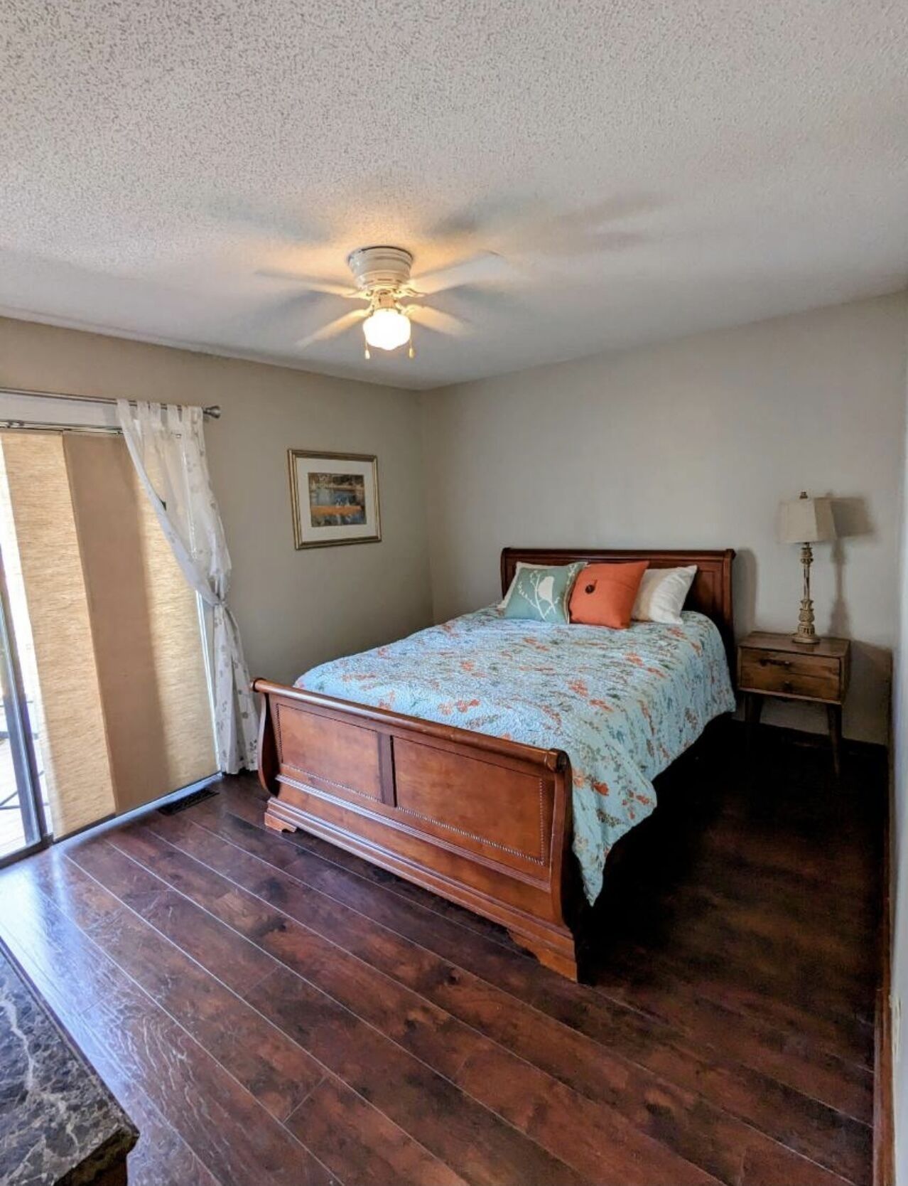 Une chambre à coucher avec un lit en bois, un ventilateur de plafond, une table de chevet avec une lampe et une fenêtre avec des rideaux.