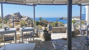 Une terrasse extérieure avec un sol en pierre, des tables et des chaises blanches, et une vue sur la mer et le paysage rocheux.