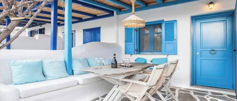 A room with a white sofa, blue shutters, a table with glasses and a bottle, and a stone floor.