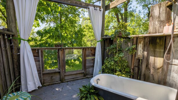 Una terraza de madera con una bañera, cortinas blancas y plantas en macetas.
