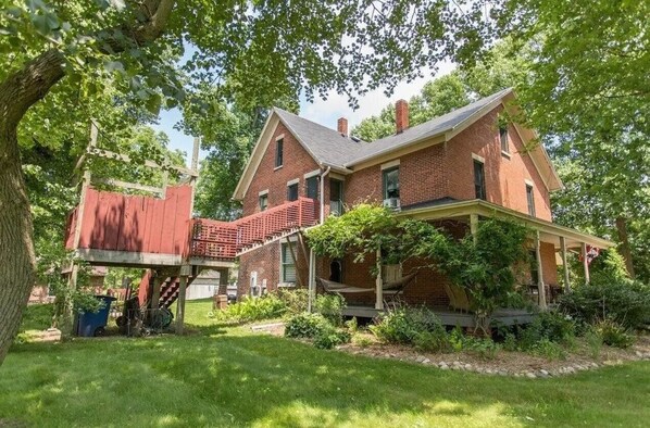 A two-story brick house with a covered porch and a small red structure attached to the left. - Historic Farmhouse with Hot Tub, Treehouse Shower, & Chickens! (Michigan City)
