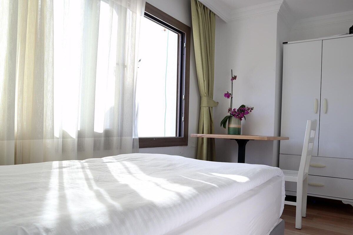 Un dormitorio con cama, una ventana con cortinas translúcidas, una mesita con un florero y un armario blanco.