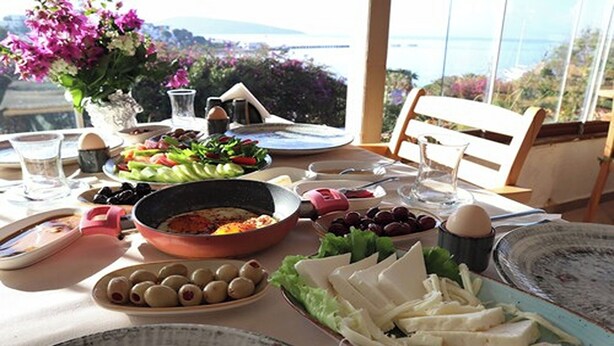 A table set with various dishes, including olives, cheese, and a salad, with a scenic view in the background. - DÖRTMEVSİM PANSİYON (Bodrum)