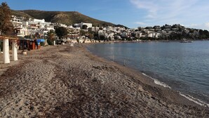 A coastal town with buildings along the shoreline, a pebbly beach, and a clear sky. - DÖRTMEVSİM PANSİYON (Bodrum)