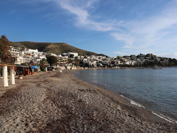 A coastal town with buildings along the shoreline, a pebbly beach, and a clear sky. - DÖRTMEVSİM PANSİYON (Bodrum)