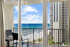 A balcony with a view of the beach and ocean, a chair, and a building with balconies.