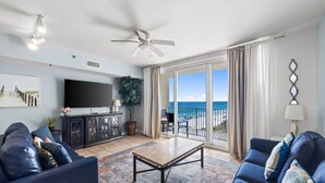 A living room with a flat-screen TV, dark blue sofas, a coffee table, and a view of the ocean through large windows. - Ocean view condo w/ pool and hot tub (Panama City Beach)