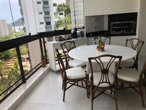 A balcony with a round table and chairs, a small kitchen area with a grill, and a view of buildings and greenery. - M3 Kingston Town 61 (Bertioga)