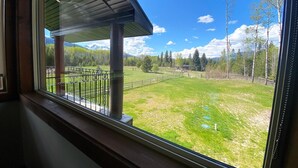 A view from a window overlooking a grassy area with a metal fence, a wooden deck, and a mountain range in the background. - Westview Cabin Rental (Valemount)
