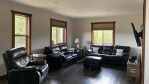 A spacious living room with a large black leather sofa, a smaller leather armchair, and a wooden coffee table. There are two windows providing natural light, and a television mounted on the wall. - Westview Cabin Rental (Valemount)