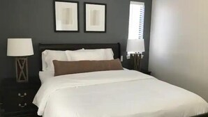 Un lit bien fait, avec du linge de lit blanc et un coussin décoratif marron, flanqué de deux tables de chevet avec des lampes, dans une pièce aux murs sombres et où deux tableaux encadrés sont accrochés au-dessus du lit.