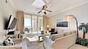 A hotel room with a sofa, armchair, TV, ceiling fan, and a view of the city. - Azure 319 - Beachfront Condo w/ Coastline Views - Free Seasonal Beach Service (Fort Walton Beach,)