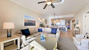 A modern living room with a sectional sofa, a coffee table, and a ceiling fan. There is a kitchen area visible in the background. - Azure 319 - Beachfront Condo w/ Coastline Views - Free Seasonal Beach Service (Fort Walton Beach,)