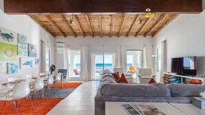 A modern living room with a wooden ceiling, a grey sofa, a white coffee table, and a TV. There are white chairs and a dining table with orange chairs.