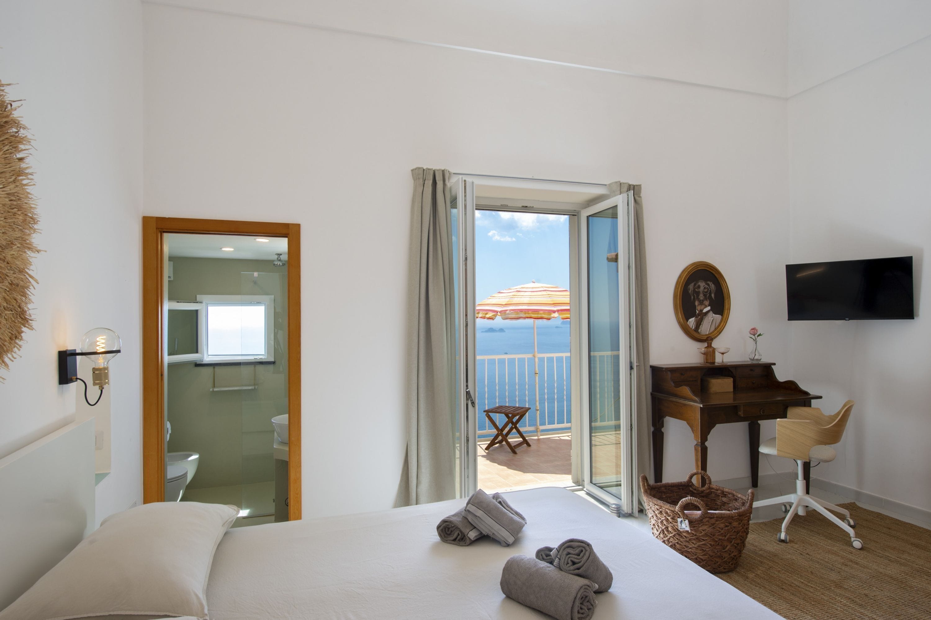 A hotel room with a bed, a desk, a chair, a TV, and a balcony with a view of the sea.