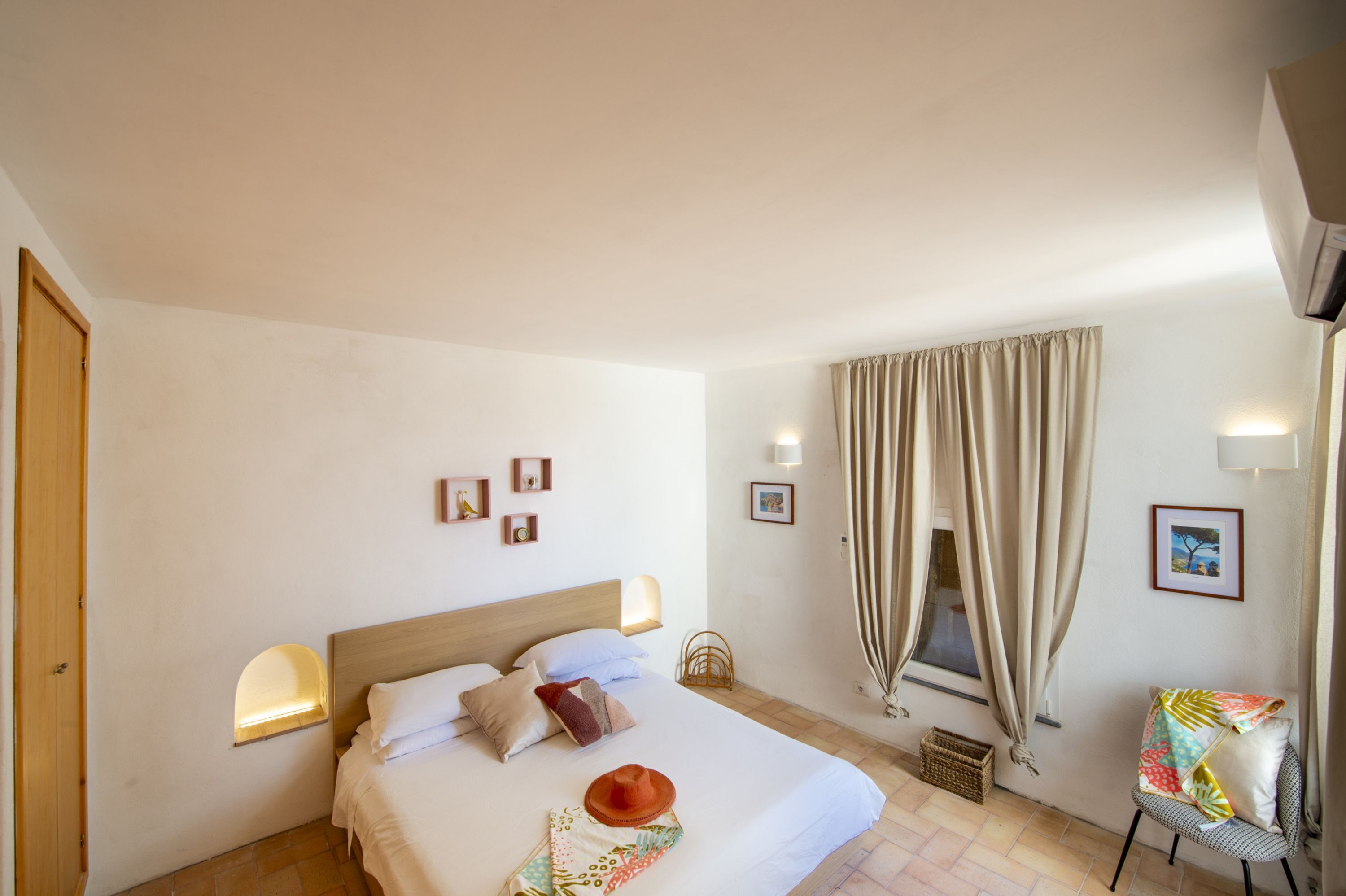 Un dormitorio con cama, una silla, una ventana con cortinas e iluminación en la pared.