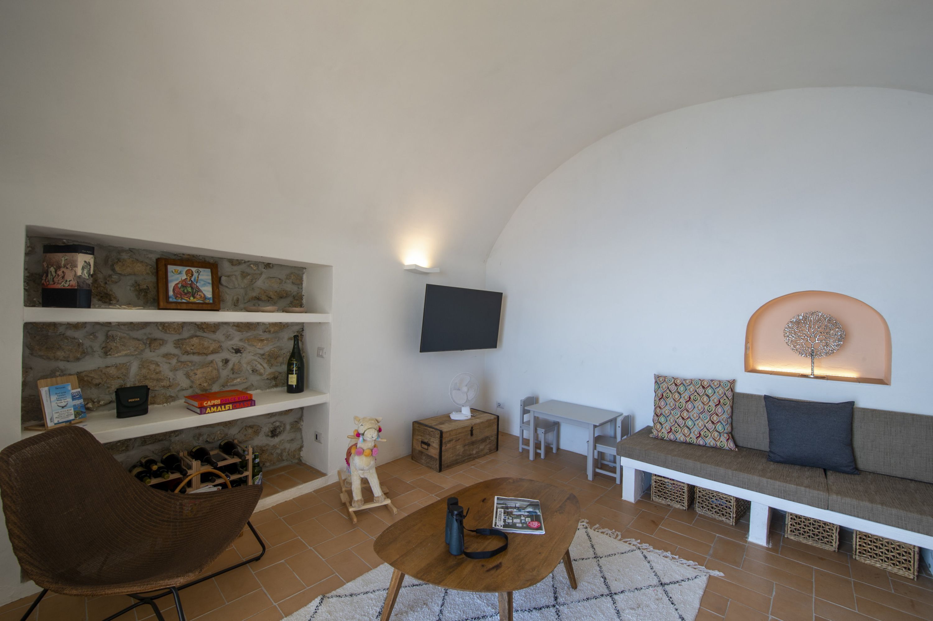 Un salon chaleureux avec un mur en pierre, une table basse en bois, un fauteuil en osier, un canapé avec des coussins et un téléviseur fixé au mur.