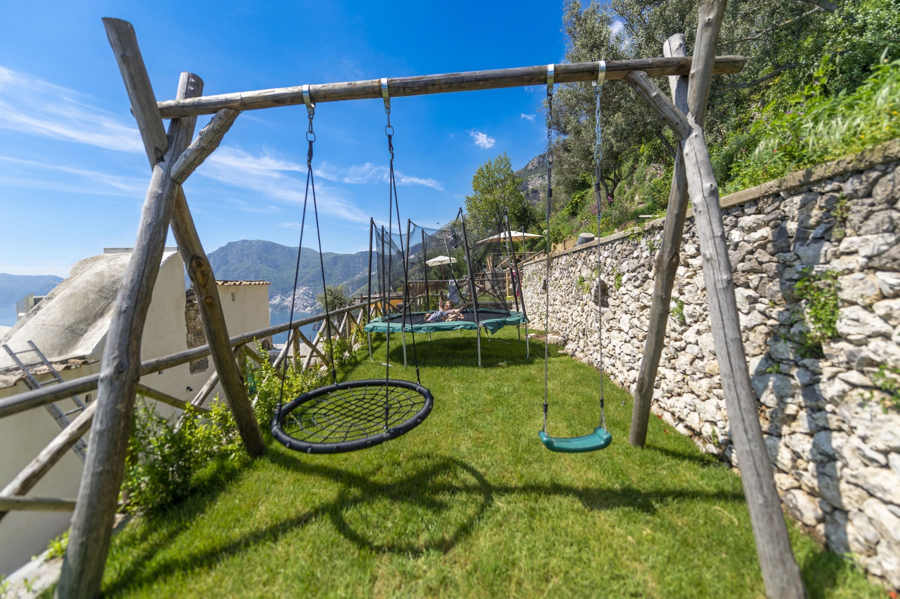 A wooden swing set with a netted swing, a trampoline, and a stone wall.