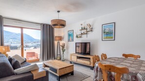 A modern living room with a sofa, coffee table, TV, dining table, and chairs. There is a view of mountains through the window.