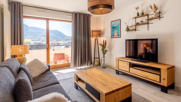 A modern living room with a sofa, coffee table, TV, and a view of mountains through large windows.