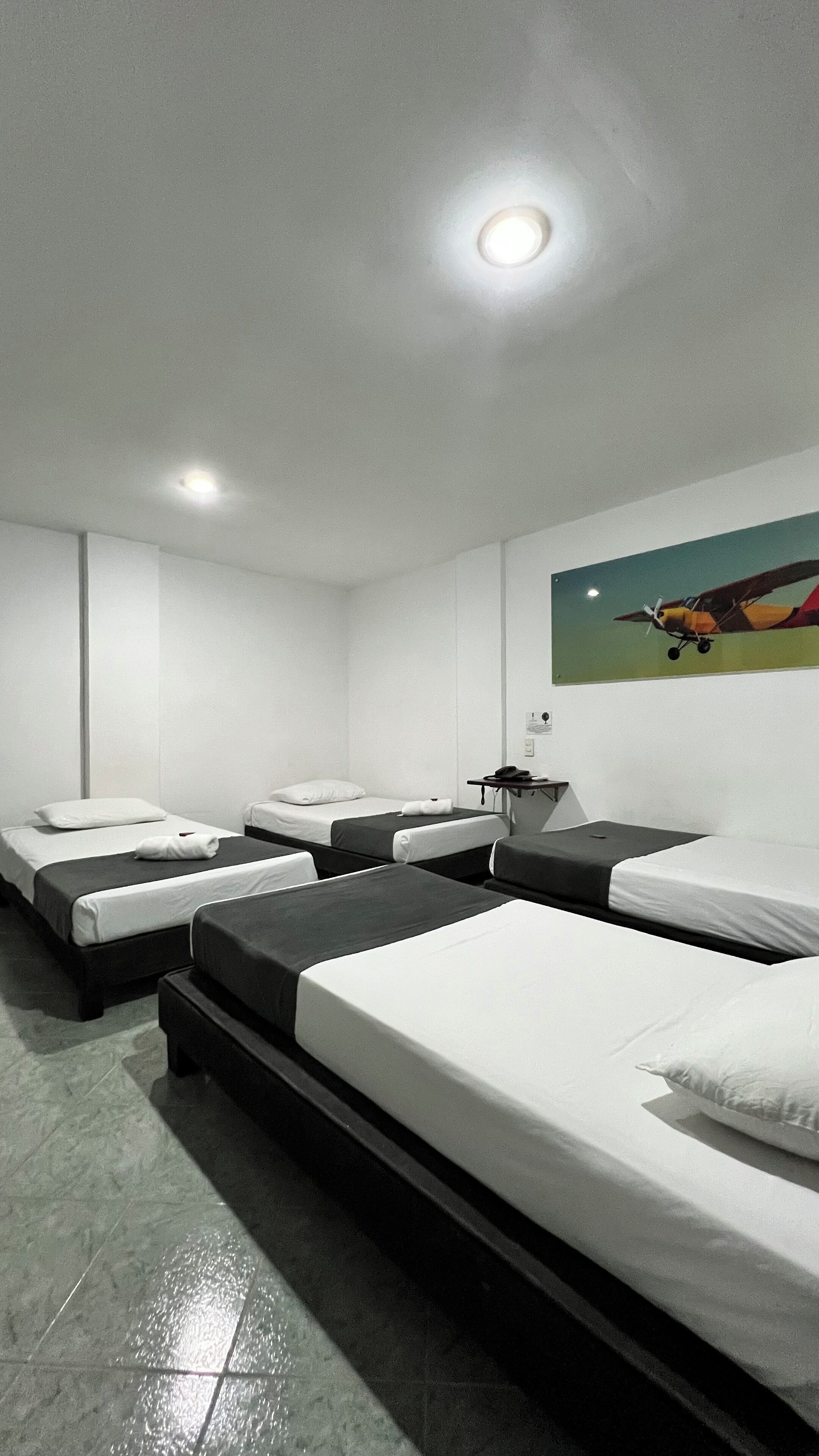 Ein Hotelzimmer mit vier Einzelbetten, einem kleinen Schreibtisch mit Telefon und einem Bild von einem Flugzeug an der Wand.