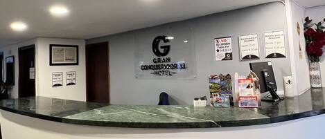 A hotel reception desk with a glass partition, a sign for 'Gran Conquistador 33 Hotel', and a 'Tour a la Casa' brochure.