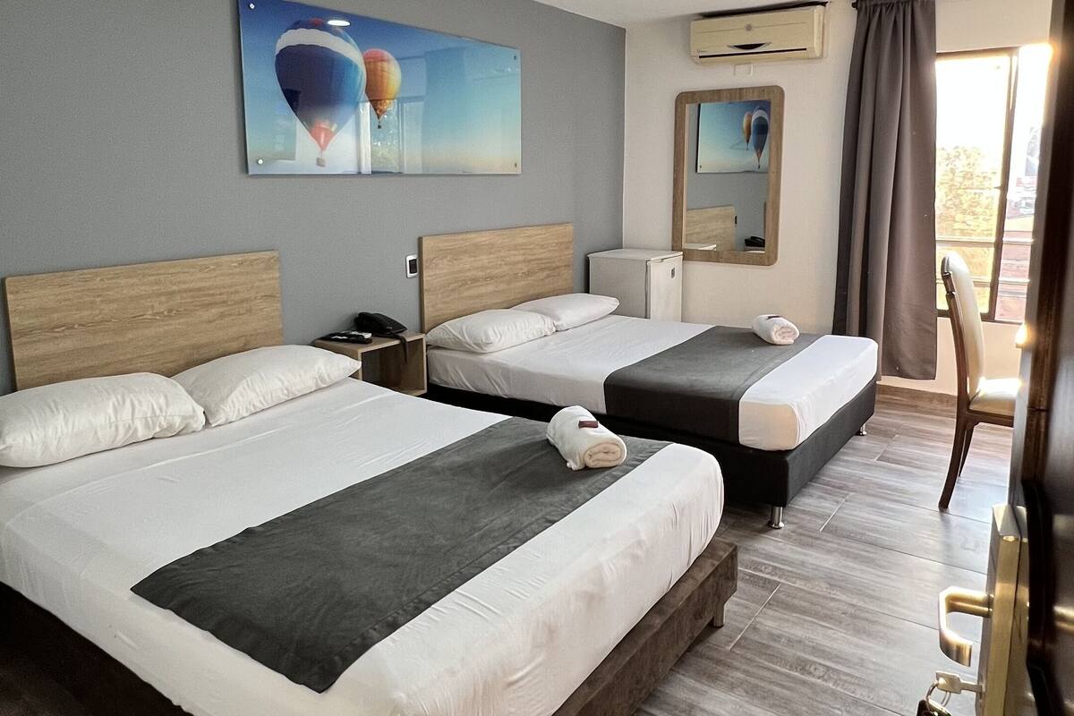 Een hotelkamer met twee bedden, een klein bureau, een stoel en een groot raam met gordijnen.