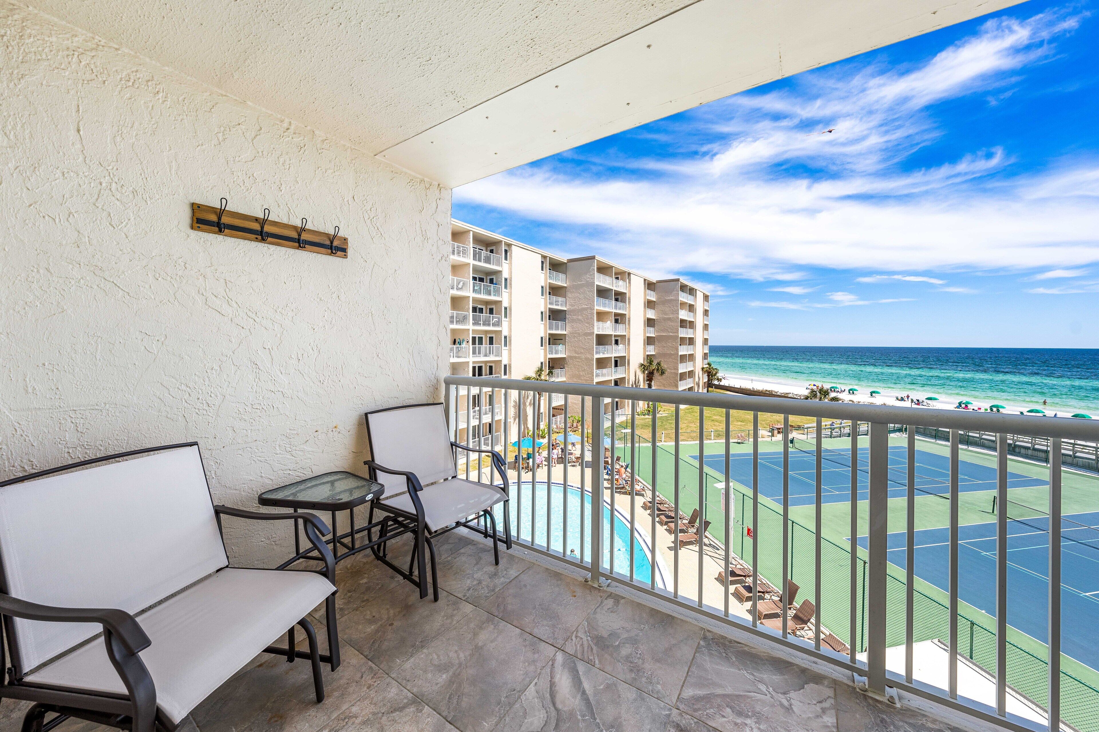 A balcony with a view of the ocean, a tennis court, and a pool area.