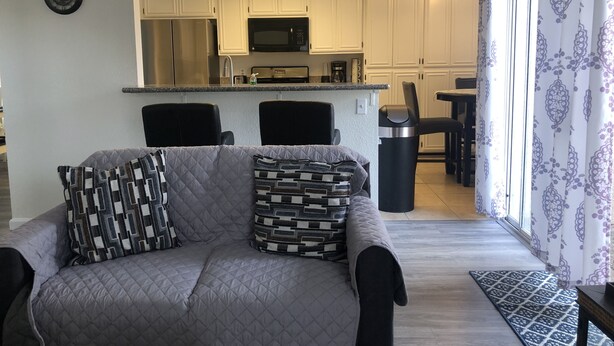 Una sala de estar con sofá gris, cojines estampados, una mesita de centro y vista a una cocina con armarios blancos y microondas.