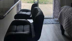 Una habitación con puerta corrediza de vidrio que da a un patio, un sofá negro, una cama con cubrecolcha acolchado y una pequeña mesa con una silla.